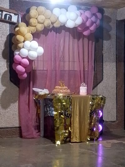 The image shows a party setup in what appears to be Aricagua, Venezuela. The background consists of a window covered with a rose-colored curtain. A balloon arch, colored gold, white, and pink, is draped around the window. In front of the curtain is a small table, draped with gold and shiny materials. On the table are a cake and a decorative item, possibly a candle holder. To the right side of the table, there's an object emitting a purple glow. The floor is a smooth surface, possibly concrete. The environment seems to be indoors, against a wall. There are no people visible in the image.