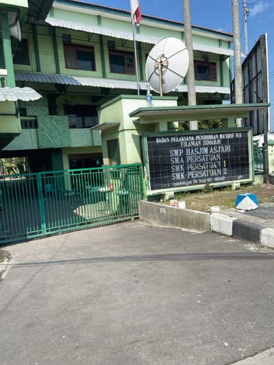 The scene is a school entrance in Kecamatan Tulangan, Indonesia. The school is a two-story building with a green facade and a satellite dish on the roof. The entrance to the school is through a green metal gate. There is a sign on the right side of the gate that reads "BADAN PELAKSANA PENDIDIKAN MA'ARIF NU TULANGAN SIDOARJO SMP HASJIM ASJARI SMA PERSATUAN 1 SMK PERSATUAN 2". There are also several paint cans on the ground next to the sign. The ground is paved with asphalt. The sky is blue with white clouds. The location is approximately -7.48 latitude, 112.644 longitude.