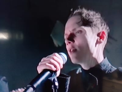 A close-up, angled, medium shot shows a young man with short, curly, light brown hair singing into a microphone. His eyes are closed, and his mouth is open in mid-song. He is wearing a dark jacket with a textured collar. The background is dark and appears to be an indoor venue. There are no clear indications of the specific location within Kempten (Allgäu), Germany.