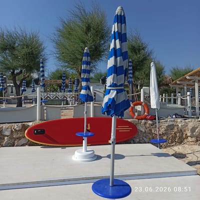 The image depicts an outdoor scene, likely a beach or resort area, in Molfetta, Italy, on June 23, 2026, at 8:51 AM. In the foreground, a bright red stand-up paddleboard rests on a concrete platform. Several closed beach umbrellas are visible, predominantly with blue and white stripes, along with one white umbrella. The umbrellas are supported by metallic poles with blue bases. Behind the umbrellas, a low stone wall separates the platform from a sandy area. Beyond the wall, a white fence with a rope railing encloses a section of the area. Several trees with sparse, green foliage are scattered throughout the scene. In the background, there are some small structures, possibly changing rooms or concessions, with thatched roofs. The sky is clear and blue. No people are visible in the image.