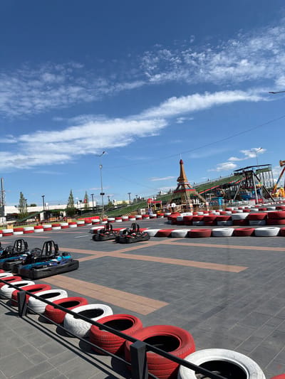 The image captures a sunny day at a go-kart track in Tohatin, Moldova. The track is bordered by stacks of red and white tires, forming a barrier. Several go-karts are parked on the track, with two in the foreground clearly numbered '1' and '2'. In the background, an amusement park area is visible, featuring a replica of the Eiffel Tower, a roller coaster, and other carnival rides, set against a bright blue sky with scattered white clouds. The ground is paved with dark grey interlocking bricks. The scene is devoid of people actively participating in go-karting or on the rides, suggesting it might be before opening hours or during a lull.