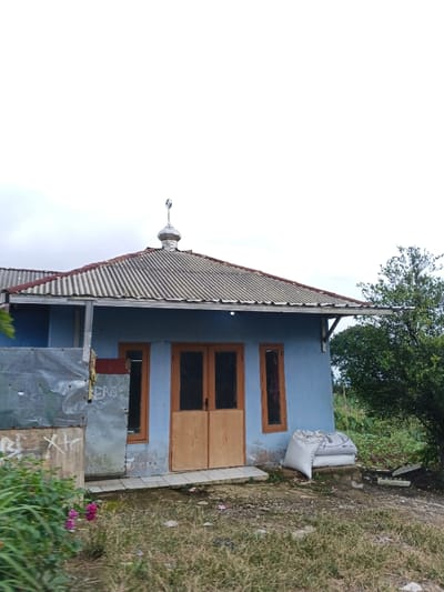 A single-story structure with light blue walls and a corrugated metal roof is depicted under an overcast sky, located in Kabiufa, Papua New Guinea. A silver-colored dome finial, topped with a crescent moon and star symbol, is mounted centrally on the roof ridge. The building's facade features a central set of light brown wooden double doors with dark handles, flanked by two narrow, tall windows with matching light brown frames. A shallow concrete step fronts the entrance. To the left of the entrance, an improvised barrier of corrugated metal sheets, marked with graffiti including "XTC" and "ERIEB," partially obscures the building's side. A concrete block is visible in front of this barrier. In the foreground, the ground is covered with dry grass and sparse green vegetation, including small pink flowers on the far left. Two large white sacks are stacked against the building's right side, adjacent to a small tree with green foliage. No individuals are present.