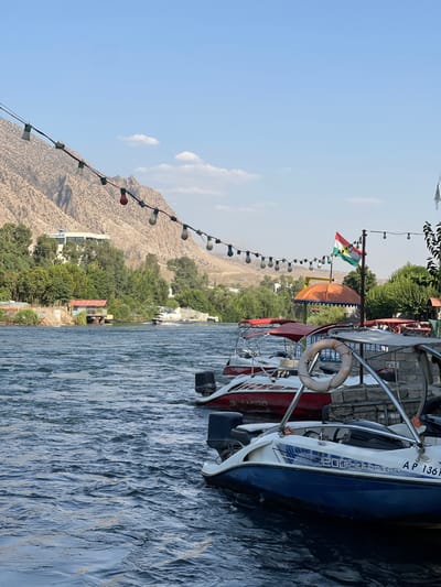The image captures a vibrant daytime scene at a waterfront location, likely a lake or river, set against a backdrop of mountains. The perspective is from the water or a very low vantage point on the bank, looking towards the shore and mountains.
In the foreground and extending to the mid-ground on the right side, several boats are moored or docked. The closest boat is a white and blue motorboat or speed boat. It features a white hull with a distinct blue stripe on its upper side. Visible on its side are the words "20C-FEESTER" and the registration number "AP 1361". A white lifebuoy hangs from its railing, and an outboard motor is partially visible at its stern. Behind it, another boat is predominantly red, with "SEA DOO" visible on its side. Further back, additional boats with canopies or covers are visible. The water immediately surrounding the boats and stretching across the lower half of the image is choppy with ripples, reflecting the clear daylight.
Stretching diagonally from the upper left corner down towards the center of the image is a string of outdoor lights, with multiple bulbs, some appearing clear/white and at least one red, strung across the scene, likely over a promenade or seating area. Above the boats and string lights, a flag is flying from a pole. This flag displays horizontal stripes of red, white, and green, with a prominent golden sun emblem in the center, identifiable as the flag of Iraqi Kurdistan (Kurdistan Region).
Beyond the water and the immediate shoreline, a lush band of green trees and vegetation covers the banks, interspersed with what appear to be small buildings or structures. In the distant background, towering, rugged mountains with a dry, earthy, light brown hue dominate the horizon. These mountains show sparse vegetation. The sky above is a clear, bright blue with a few scattered, wispy white clouds. The overall impression is that of a sunny day at a popular leisure or tourist spot by the water, surrounded by natural beauty.