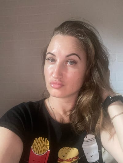 A medium close-up, selfie-style photograph shows a woman with medium-toned skin, light brown hair, and hazel eyes. She is wearing a black t-shirt with a graphic design of french fries, a hamburger, and a milkshake. She has a small nose ring and is wearing a silver necklace and a wrist watch. The background appears to be a brick wall, painted a neutral color. The lighting is soft, and the image has a slightly grainy texture. The location is not specified.