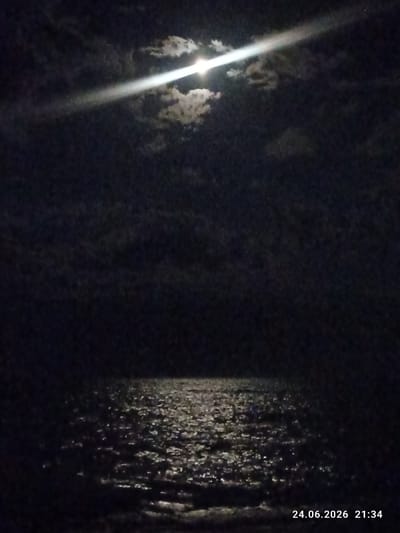 The image depicts a night scene over a body of water, likely the Caspian Sea near Shybartubek, Kazakhstan. The moon is partially obscured by clouds, casting a bright beam of light across the sky and reflecting on the rippling water. The moon's luminescence creates a shimmering path on the dark surface, highlighting the texture of the waves. The overall atmosphere is serene and somewhat mysterious, with the dark sky and the solitary beam of moonlight dominating the composition. The presence of clouds adds depth and texture to the night sky. A timestamp in the bottom right corner reads "24.06.2026 21:34," indicating the time and date the photograph was taken. There are no people or man-made objects visible in the frame, emphasizing the natural beauty of the night landscape.