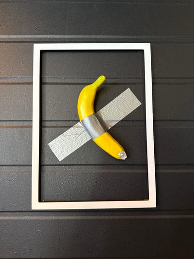 A single, ripe banana is affixed to a dark, textured wall with silver duct tape. The banana is positioned diagonally within a simple, white rectangular frame. The frame is empty and does not contain a backing or glass. The wall appears to be made of horizontally oriented panels, with a dark grey, speckled surface. The lighting suggests an indoor setting. There are no people or any other objects visible in the scene. The image is a photograph taken from a straight-on perspective, with the banana and frame centered.