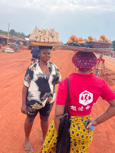 The image depicts a bustling street scene in Otukpa, Nigeria. Two women are prominently featured, each carrying goods on their heads. The woman in the foreground wears a black and white floral shirt and shorts, balancing a tray piled high with what appear to be nuts or seeds. The second woman, behind her, is dressed in a red t-shirt with a white logo, a patterned yellow and blue skirt, and a polka-dot headscarf. She also has a cell phone in her hand and carries a tray on her head, filled with round, orange-colored fruits. The ground is a reddish-brown color, indicative of the local soil. In the background, other figures and vehicles can be seen, suggesting a busy market or commercial area. The sky is overcast.