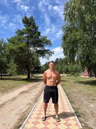 A shirtless man with short, dark hair and a muscular build stands facing the camera outdoors. He is wearing black shorts and olive-green slide sandals. The man is standing on a decorative paved path with a pattern of yellow and pink diamond shapes. To his left, a sandy path runs parallel to the paved path. The background features a grassy area and numerous trees, including tall pine trees and leafy deciduous trees. In the distance, there are hints of buildings and structures, suggesting a park or recreational area. The sky above is blue with scattered white clouds. The scene appears to be in a natural setting, possibly a park in the city of Lebyazhye, Russia, during daytime.