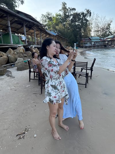 In this vertical, medium-range shot taken on a sandy beach, two women stand near the water's edge, one of whom is holding a smartphone to take a selfie. The woman on the left, wearing a floral print dress, is looking at the phone. The woman on the right, dressed in a light blue and white checkered dress, is partially visible behind her, also facing the phone. In the background, there are several wooden structures, possibly huts or restaurants, built on stilts along the coastline. Rocky formations are visible on the left side of the image, and in the distance, trees and a calm body of water with gentle waves can be seen. The sand is light brown and appears damp in some areas. Another woman with glasses is seated on a chair in the middle ground, facing away from the camera. Several empty wooden chairs and tables are scattered on the beach. The sky is clear and overcast, suggesting it is either early morning or late afternoon. The overall scene depicts a relaxed atmosphere at a beachside location. The specific location in Thuận Thành, Vietnam, is not identifiable from the visual cues in the image.