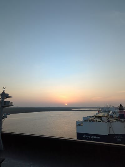 The image shows a wide shot of a harbor at sunrise in Hambantota, Sri Lanka. The sky is a gradient of pale blue and orange. In the distance, the sun is visible as a bright orb on the horizon, casting a golden light across the water. A large cargo ship, identified by the text "VENUS LEADER" on its side, is docked in the harbor. The ship is white with a dark blue hull and has several decks and structures. To the left of the image, part of another vessel or structure, possibly a port crane, is visible. The water is calm and reflects the colors of the sky. The shoreline in the distance appears to be a flat, dark landscape. The foreground shows a dark, solid surface, likely the deck of a ship or a pier, with a railing. There are no people visible in the image. The overall scene depicts a tranquil dawn at a Sri Lankan port.