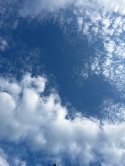 The image depicts a clear blue sky with scattered white clouds. The clouds are varied in size and density, with some appearing as wispy streaks and others as larger, fluffier formations. The sunlight is diffused through the clouds, creating a bright and airy atmosphere. The bottom of the frame shows a very small portion of what appears to be the tops of trees or vegetation, suggesting an outdoor environment in Angarskie Khutora, Russia. No people or specific structures are visible in the image.