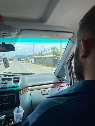 The image, taken from inside a car, depicts a forward view of a road in the city of Khalimbekaul, Russia. The driver's right shoulder and head are visible in the foreground, with a portion of the car's interior, including the dashboard, air conditioning vents, and a car stereo system displaying "Bluetooth 5.1," also visible. A plastic cup with a straw is placed in the center console. Outside the car, the road stretches forward, with buildings and fences lining the sides. Several vehicles, including a white van, are seen on the road. Power lines and utility poles are also present. The sky is partly cloudy, and a mountain range is visible in the distance. A sun visor with an "AIRBAG" warning label is positioned at the top of the windshield. A phone with an eagle design is hanging from the rearview mirror.