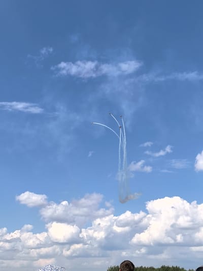 The image depicts an outdoor event under a clear blue sky dotted with fluffy white clouds. In the upper half of the frame, three small planes are performing an aerial maneuver, leaving trails of white smoke against the sky. One plane is slightly higher and to the left, while the other two are positioned below and to the right, their smoke trails converging and forming a downward, loop-like shape. In the lower portion of the image, a crowd of people are visible from behind, suggesting they are spectators. The foreground is partially obscured by the heads of individuals and a patterned fabric item. A line of trees forms a distant horizon beneath the sky. The scene is likely a public gathering, possibly an airshow, taking place in or near Ustinovo, Russia.