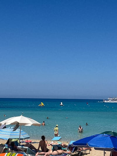 The image depicts a sunny day at a beach in Protaras, Cyprus, with clear blue skies and calm turquoise waters. In the foreground, beach chairs and umbrellas are set up on the sandy shore, with several people relaxing on the chairs. Some are sunbathing, while others are partially obscured by the umbrellas.
In the water, numerous people are swimming and wading. A yellow pedal boat with occupants is visible further out. In the background, a large white boat, likely a cruise ship or ferry, is sailing on the horizon, with "enjoy Coca-Cola" visible on its side. The scene is characteristic of a popular tourist destination, with a lively atmosphere of people enjoying the seaside.