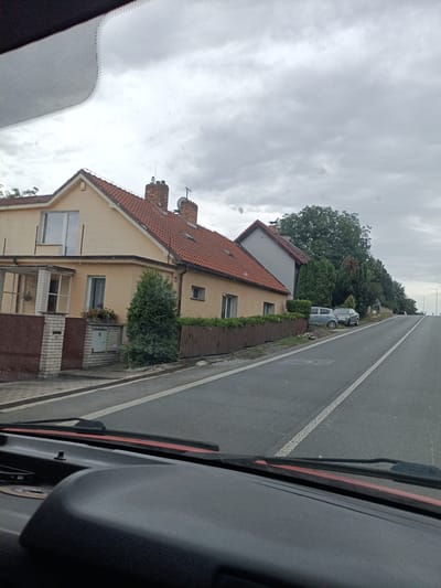 The image captures a residential street scene in Cvrčovice, Czechia, under an overcast sky. The perspective is from inside a vehicle, looking out through the windshield. On the left side of the road, there are several houses with red tile roofs. The house closest to the foreground is a two-story building with a light yellow facade, featuring a covered porch area with glass panels. A brick fence and a brown gate are visible in front of this house. Further down the street, another house with a gray facade and a red roof is visible. A wooden fence runs along the property line of these houses. Two cars, a light gray hatchback and a dark gray sedan, are parked on the side of the road near the houses.
The road itself is paved and has white lane markings. It appears to be a gentle upward slope towards the right side of the image. Several streetlights are positioned along the right side of the road in the distance. Lush green trees line the right side of the street behind the parked cars and continue into the distance, creating a natural backdrop. The overall atmosphere is subdued due to the cloudy weather.