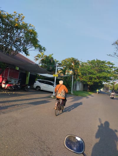 In Sidodadi, Indonesia, the image captures a sunny street scene. The foreground is dominated by the perspective of a rider on a motorcycle, with their mirror visible on the lower right. Directly ahead, an elderly person is cycling away from the camera, wearing an orange patterned shirt and dark pants, with a bicycle basket on the back. Their shadow stretches across the asphalt road. To the left of the road, a building with a dark canopy and red accents is visible, with two scooters parked in front of it. A white van is parked to the right of the building. Further down the road, a person in a yellow shirt walks, and in the distance, another motorcycle with two riders is visible. The environment includes trees, palm trees, a concrete wall, and clear blue sky. A yellow sign with the text "PARKIR GRATIS" (Free Parking) is mounted on a pole near the building.