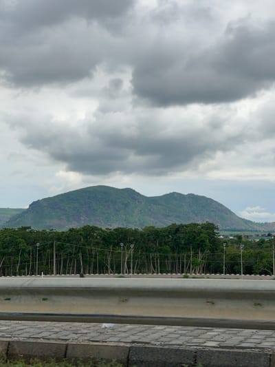 A distant, lush green mountain range with rocky outcrops dominates the background under a cloudy sky. In the mid-ground, a dense line of tall green trees forms a natural barrier. In front of the trees, a row of modern streetlights stands. A concrete barrier runs horizontally across the foreground, separating the viewer from a paved area and a grassy verge. The overall scene depicts a natural landscape near a developed area, likely on the outskirts of a Nigerian city like Immigration Quarters.