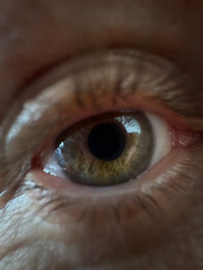 The image shows a close-up of a human eye with hazel or green iris and a black pupil. The eye is surrounded by skin, eyelashes, and a small amount of moisture at the inner and outer corners. The image appears to be taken in a well-lit environment, as evidenced by the reflections in the iris. There is no indication of the city of Ketoshi, Russia in the image.
