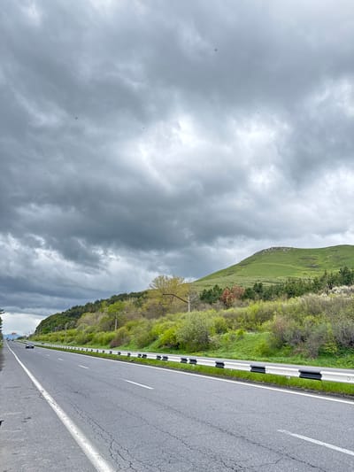 The image depicts a road scene in Tsamakaberd, Armenia. The road stretches into the distance, marked by white lines and a black-and-white guardrail on the right side. Lush green hills covered with vegetation rise on the right side of the road. Dominating the left side of the image are dark, ominous clouds. The sky is overcast, suggesting an impending storm or recent rainfall. There is a single tree present. The landscape indicates a rural or mountainous region.