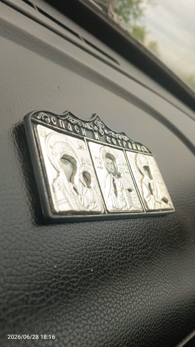 A close-up, low-angle shot captures a religious icon with a metallic silver finish, positioned on a dark, textured surface, likely the dashboard of a vehicle. The icon is embossed with intricate designs and features three panels depicting religious figures. The leftmost panel shows the Virgin Mary, the central panel depicts Jesus Christ, and the rightmost panel portrays a saint. Cyrillic text is visible at the top of the icon. The background shows a blurred view of the sky and trees through a windshield. The timestamp "2026/06/28 18:16" is overlaid in the lower-left corner, suggesting the image was taken in Luzа, Russia.