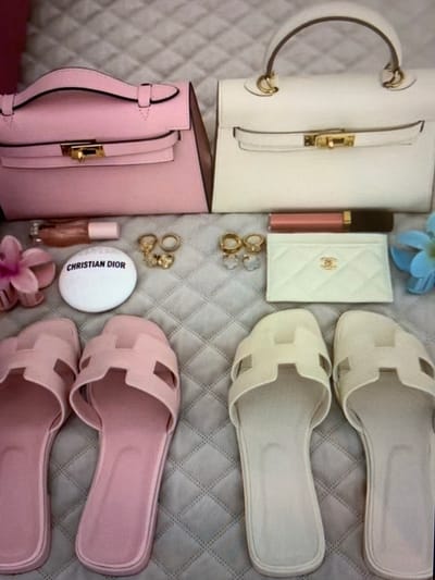 This image displays a flat lay arrangement of luxury fashion accessories. In the background, a quilted surface serves as the setting. The arrangement includes two handbags: one pink and one off-white, both with top handles and gold hardware closures. Adjacent to the handbags are several smaller items. There is a tube of lip gloss and a white circular compact with "CHRISTIAN DIOR" printed on it. A pink artificial flower is also visible. A white cardholder with a quilted pattern and a gold "CC" logo, indicative of Chanel, is present. A selection of gold-toned rings and earrings, including some with clear gemstone accents, are also arranged. Finally, there are two pairs of slides: one pair is pink, and the other is off-white. The image does not provide any information about the city of Konstantinivka, Kyrgyzstan, or any specific location within it.