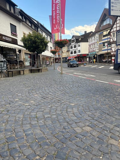 The image depicts a street scene in Adenau, Germany, with a focus on a cobblestone street that curves towards the right. The street is lined with traditional German half-timbered buildings. On the left, a white building with dark trim features awnings and signage for "AUSZEIT" (meaning "time out"). A small tree with green foliage stands in front of this building. Red and pink flowers in planters are visible.

In the mid-ground, a black car is driving on the asphalt road. Further down the street, there are more buildings, including one with "FABRY" signage. People are seated at outdoor tables at what appears to be a cafe or restaurant. A blue bus stop sign is present on the right side of the image. The sky is blue with scattered white clouds. The overall impression is of a charming, historic European town square.