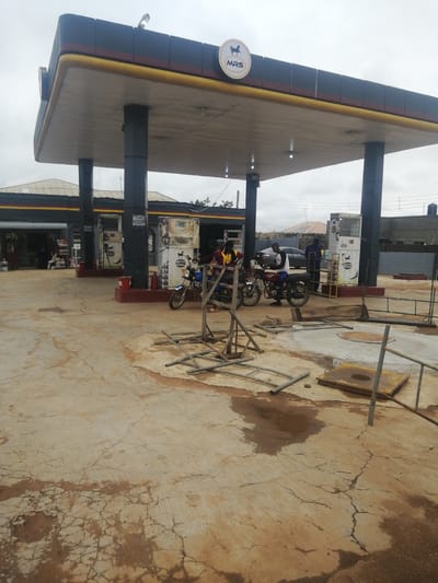 This image captures an outdoor scene at an MRS fuel station in Ajasse Ipo, Nigeria. The fuel station features a large covered canopy supported by dark gray pillars. The canopy has a dark blue exterior with red and yellow trim. A circular logo with a horse and the letters "MRS" is visible on the canopy.

Beneath the canopy, several fuel pumps are present. One pump, primarily white with black and red accents, has the MRS logo. To the left of this pump, a red bucket and a red fire extinguisher are visible on the ground.

The ground surface is made of cracked and stained concrete, with visible patches of moisture and dirt. In the foreground, a partially constructed metal scaffolding or frame lies on the ground.

Several individuals are present in the scene. Two people are seated on a red motorcycle parked near a fuel pump. Another person, wearing a white shirt with black markings and dark pants, is seated on a red motorcycle, seemingly waiting. In the background, another person in a blue outfit stands near a fuel pump. A dark-colored car is parked further in the background, and to the far left, a small shop with shelves of goods is visible through an open doorway. The sky is overcast.