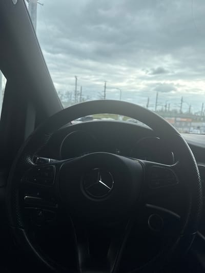 The image shows the interior of a car, focusing on the steering wheel with a Mercedes-Benz logo. The steering wheel is black with controls on either side. Outside the car window, the scene is blurry, suggesting an overcast day with utility poles visible in the background. The location is potentially the city of Amstetten, Austria, but without more specific details in the image, it is hard to confirm and identify a precise location.