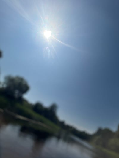 This is a vertical, outdoor, wide-angle photograph taken during the daytime, featuring a bright sun with prominent rays radiating across a clear blue sky. In the upper portion of the frame, the sun is partially obscured by lens flare, creating a glowing effect. The lower half of the image is significantly out of focus, showing a body of water reflecting the sky and blurred greenery. On the left side of the water, a sloped embankment is visible, covered in vegetation and trees. The overall scene suggests a natural landscape, possibly a river or lake, with the sun high in the sky. The image was captured in the vicinity of Kurmene, Latvia, judging by the geotag information.