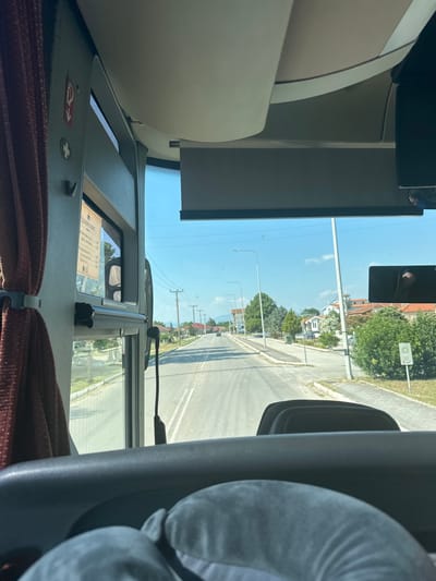 The image is taken from the interior of a bus, looking forward through the windshield. The bus is traveling down a paved road lined with trees and buildings. The sky is blue with scattered clouds. On the left side of the image, a maroon curtain is visible, as well as a sign with red text and symbols. A menu in Cyrillic script is displayed on the bus's interior wall. The road appears to be in a rural or suburban area, possibly Chaidefto, Greece, based on context. There are streetlights and power lines visible along the road. The dashboard of the bus is in the foreground, with a gray neck pillow obscuring part of it.