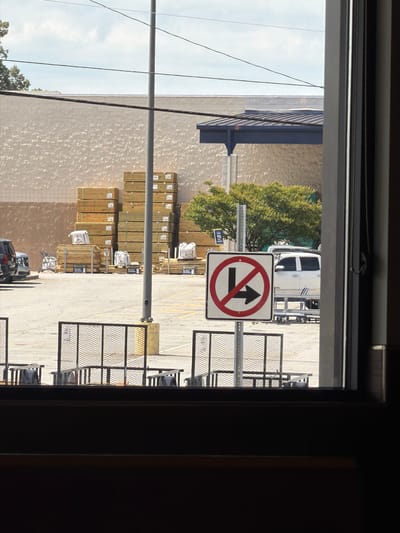 The image shows a view from inside a building looking out into a parking lot. In the foreground, a portion of a window frame is visible on the right. Beyond the window, a paved parking lot is visible with several metal safety barriers. Further back, stacks of lumber are piled up outside a large, light-colored building with a textured facade. To the right of the lumber, a dark roofline of another building is visible, supported by columns. A light-colored truck is parked in the lot. A traffic sign with a red circle and a diagonal line, indicating no left turn and no right turn, is prominently displayed in the middle ground. The sky is partly cloudy. The location is Easley, United States of America.