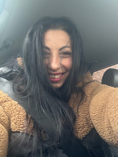 A woman with long, dark hair smiles towards the camera, partially obscured by her hair. She appears to be wearing a brown, textured jacket and a black top or scarf. The setting appears to be inside a car, indicated by the car interior elements visible. The location within Warrington, United Kingdom, is not discernible from the image.