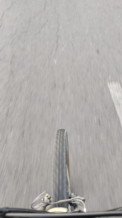 The image shows a close-up, low-angle perspective of a bicycle wheel in motion on a paved road. The road surface is gray with visible cracks and a white line marking on the right side. The motion blur indicates the bicycle is moving forward at a considerable speed. The bicycle's front wheel and part of the handlebars are visible at the bottom of the frame. The scene is consistent with a view from a bicycle traveling within Sendangrejo, Indonesia, where such roads are common.