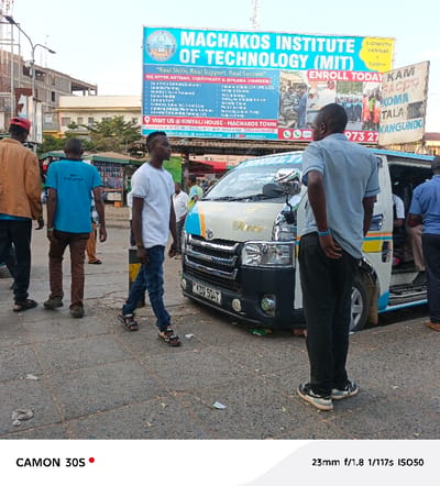 The image captures a busy street scene in Machakos Central ward, Kenya, likely depicting a public transport hub or market area. Several people are visible, mostly men, engaged in various activities related to daily commutes.
In the foreground, a man in a white t-shirt and blue jeans walks towards the right, while another man in a light blue collared shirt and dark trousers stands facing away, observing something to his right. To the left, two other men are seen from behind; one wears a brown jacket, and another sports a bright blue t-shirt with "CREATIVE" partially visible on the back.
The central focus includes a white Toyota Hiace minibus, commonly known as a matatu, with distinctive blue and yellow stripes and "Greco" written on its side. Its license plate, KDD 504T, is clearly legible. The matatu appears to be either parked or moving slowly, suggesting it's either picking up or dropping off passengers.
Dominating the background is a large billboard for the "MACHAKOS INSTITUTE OF TECHNOLOGY (MIT)", prominently displaying "Real Skills, Real Support, Real Success". It advertises artisan, certificate, and diploma courses, listing various programs and providing contact information including "VISIT US @ KINYALI HOUSE - MACHAKOS TOWN" and "ENROLL TODAY". Another smaller, partially visible billboard to the right mentions "KAM TALA KANGUNDG".
The setting is an urban environment with paved ground, and buildings are visible in the distance. The soft, warm lighting suggests it is late afternoon or early evening under a clear sky. The overall atmosphere is one of routine activity in a bustling Kenyan town. The photo was taken with a CAMON 30S, at 23mm, f/1.8, 1/117s, and ISO50.