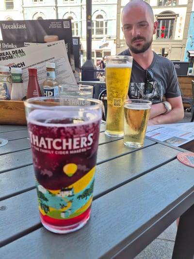 The image shows a man at an outdoor table, possibly in Cardiff, United Kingdom, based on architectural style and brand references in the scene. He is positioned with his arms resting on the table. In front of him are three beverages: two clear drinks in shorter glasses and one darker, possibly berry-flavored drink in a Thatchers-branded glass, indicating cider. Nearby are menus with breakfast and drink options. Food condiments like sauces are present. The surrounding environment consists of buildings with distinct architectural designs. The B.B.Q sign indicates an establishment nearby. The overall atmosphere is of an outdoor dining setting.