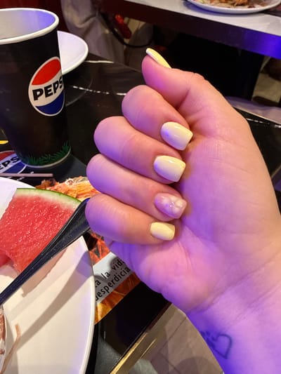 A person's hand, adorned with pastel yellow and light purple nails, is the central subject of this media file. One nail, on the ring finger, has a subtle floral design in white and pink. The hand is positioned over a collection of objects and a partially visible scene.
In the foreground, a black disposable cup with the red, white, and blue Pepsi logo is visible. Next to it, a slice of watermelon is partially submerged in a white bowl, with a black utensil resting on the rim of the bowl and partially covering the watermelon. In the background, other objects are out of focus, including what appears to be a white plate on the left and dark surfaces.
The lighting suggests an indoor setting, possibly a restaurant or cafe, with a purple hue cast over the hand, potentially from ambient lighting or a nearby light source. The overall mood conveyed by the image is casual and relaxed. The visible text is limited to the Pepsi logo on the cup. The provided location context is Carretera De San Martín, Spain, which suggests this scene might be taking place in that area. The time of day and weather are not discernible from the image.