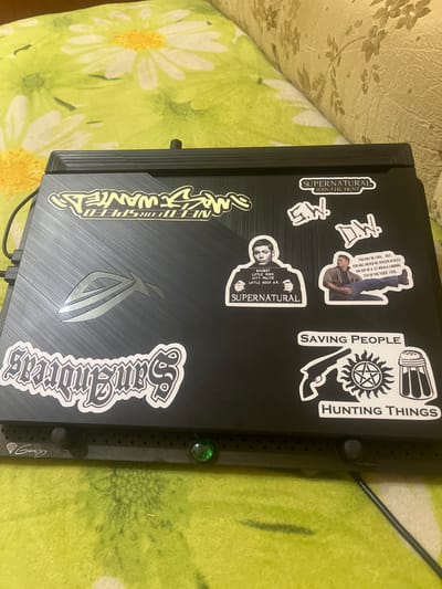 The image shows a black laptop with various stickers on its lid. These stickers include "Need for Speed: Most Wanted," "San Andreas," "Supernatural" with the tagline "Join the Hunt," and various images related to the show "Supernatural," such as mugshots of characters, quotes, and symbols. The laptop is placed on a green surface with a floral pattern, likely a blanket or tablecloth. A portion of a beige upholstered furniture item is visible in the upper background. The laptop is connected to a black cable. A green LED light is visible on the front edge of the laptop. The scene appears to be indoors, in a domestic setting. There is no identifiable information or context within the image that places it specifically in the city of Dobrotvir, Ukraine.