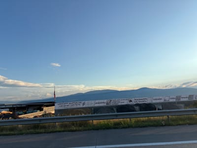 The image shows a wide, outdoor landscape scene under a clear blue sky with scattered clouds. In the foreground, a road barrier made of metal railing separates the viewer from a grassy embankment. Beyond the barrier, a construction or industrial site is visible, featuring a long, dark-roofed structure with stacks of building materials, including wood planks and possibly bagged items, arranged in neat rows.
Further back, a series of large white banners are stretched across a fence or wall. These banners display text in a non-English script, presumably Georgian, along with some graphical elements, including what appears to be a QR code on one of the banners. A Georgian flag is also visible, flying from a flagpole near the industrial site.
In the background, rolling hills and mountains form the horizon, bathed in soft sunlight. The overall impression is one of a rural or semi-industrial area, possibly on the outskirts of a town or city. The location is identified as Gorovani, Georgia. There are no people visible in the image. The lighting suggests it is either late afternoon or early morning.