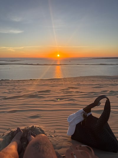 A first-person perspective shot captures a sunset over the Dune of Pilat in La Teste-de-Buch, France. The foreground is dominated by rippled sand dunes. In the mid-ground, a black woven bag is placed on the sand, with a white cap and sunglasses hanging from its strap. Two feet, one with a tattoo, are visible at the bottom left of the frame, partially buried in the sand. A pair of orange flip-flops rests in front of the bag. The background features a wide expanse of water, with the sun setting on the horizon, casting a warm orange glow across the sky and reflecting on the water's surface. The sky transitions from a pale blue at the top to a warm orange near the horizon, with thin wisps of clouds. A distant sailboat and a boat are visible on the water.