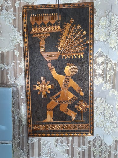 The image depicts a piece of folk art, possibly a wall hanging, crafted from straw or a similar natural material. The artwork features a stylized human figure in traditional attire, dancing or presenting a bird. The figure is depicted in motion, with one leg forward and arms outstretched. The bird, also stylized, is perched on the figure's outstretched hand, with a large, elaborate tail. The artwork is framed by a border of diamond and triangular patterns. The background of the artwork is a dark, textured material. The wall behind the artwork is covered in patterned wallpaper featuring vertical stripes and floral motifs. The overall style of the artwork suggests a connection to traditional Slavic or Eastern European crafts. The location is identified as Блеўчыцкі сельскі Савет, Belarus.