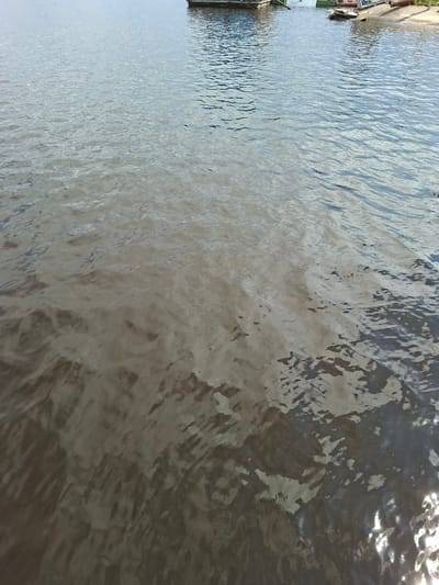 The image shows a body of water, likely a river or lake, with a distinct separation of water clarity. The upper portion of the water is a lighter, clearer blue, reflecting the sky. Towards the bottom of the image, the water becomes significantly darker and murkier, with a muddy brown appearance. This suggests a mixing of different water sources or a change in sediment load. In the background, a small portion of the shoreline is visible, featuring what appear to be boats or a dock, and some vegetation. The overall scene is a natural landscape with elements of human activity in the distance. The location, Verkhny Uslon, Russia, is a settlement on the banks of the Volga River, known for its scenic beauty and historical significance.