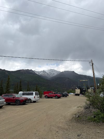 The image shows a cloudy sky over a mountainous landscape in Denali National Park and Preserve, United States. In the foreground, there is a gravel parking area with several vehicles, including a red SUV, a white van, and two red pickup trucks. Some industrial equipment and structures are visible in the background. The mountains are covered in dark green trees, with patches of snow on the higher peaks. Power lines are strung across the top of the image.