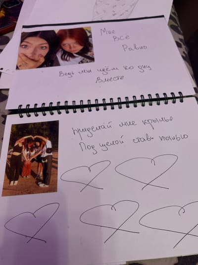 This image depicts a page in a spiral-bound notebook. On the upper half of the page, a photograph features two young women. One woman on the left has dark hair and is covering her mouth with her hand, revealing a ring on her finger. The woman on the right has reddish-brown hair and is looking towards the camera with a neutral expression. Text in Russian is handwritten next to the photograph. The text reads, "Мне всё равно" (Mne vsyo ravno), which translates to "I don't care." Below this, more text reads, "Ведь мы идём ко дню вместе" (Ved' my idyom ko dnyu vmeste), meaning "After all, we are walking towards the day together."
The lower half of the page contains another photograph, this one featuring four individuals standing outdoors, forming a heart shape with their arms. Below this photograph, more Russian text is written: "придемаи мне крылья" (pridemai mne kryl'ya), translating to "give me wings," and "под ценой ставь пылью" (pod tsenoy stav' pyl'yu), which translates to "under the price put dust." Scattered across the bottom of the page are several hand-drawn heart shapes. The notebook appears to be resting on a patterned surface, possibly a blanket or fabric. The location is not specified as Vaganovo, Russia.
