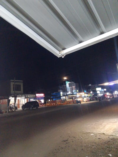 This image captures a nighttime scene in Gampongpaya, Indonesia. The foreground is dominated by a dark, asphalt road, with patches of light reflecting off its surface, suggesting artificial lighting. A metal roof structure extends from the top of the frame, casting a shadow and framing the scene.
In the background, a street is lined with various establishments, illuminated by artificial lights, indicating that it is evening or night. Visible signs include "WM BOUTIQUE" and "WM DIMSOM" with additional text "Mgudinesium Premium 2.000". The street is active with several vehicles, including a black SUV parked in front of a building, and numerous motorcycles parked and in motion. Orange chairs are arranged in front of some establishments, suggesting outdoor seating or dining areas.
The overall atmosphere is that of a bustling street at night, likely a commercial area with shops and food stalls. The lighting creates a somewhat moody ambiance, with bright lights from the shops contrasting with the dark sky. There are no discernible weather conditions mentioned, but the scene suggests a clear night. The activity captured is typical of a lively urban or semi-urban environment at night.