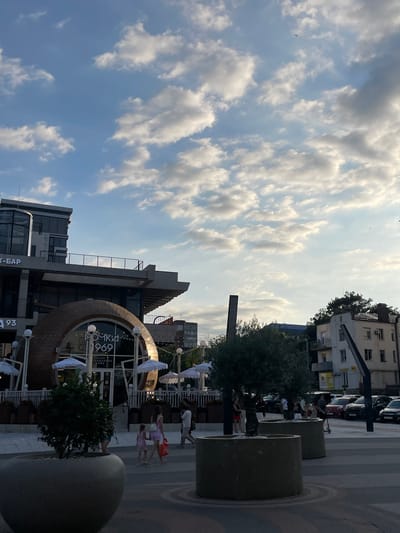 This image depicts an outdoor scene in Ivanovskaya, Russia, likely during the late afternoon or early evening, given the quality of light. The sky is a prominent feature, filled with a mix of scattered white clouds against a bright blue background, suggesting fair weather.

In the foreground, several large, cylindrical planters made of concrete are visible. Beyond these planters, a street or pedestrian area is paved with patterned tiles. The mid-ground features a building with a distinctive circular facade, partially visible through a large, arched opening. The text "ОДЯНКА 969" and "ГЕНДАРНОЕ КАФЕ" is visible on the building, indicating it is likely a cafe. White umbrellas or sunshades are set up in front of the cafe.

Several people are scattered throughout the scene. Some are walking, while others appear to be socializing or dining at the cafe. Two young girls, one in a pink dress and the other in a purple dress, are walking together. In the background, there are more buildings, including a multi-story apartment building with windows and air conditioning units. Cars are parked along the street behind the buildings.

The overall atmosphere appears relaxed and casual, suggesting a typical urban environment during a pleasant time of day. There are no strong emotional cues, but the presence of people outdoors and the setting of a cafe suggest a social or recreational activity.