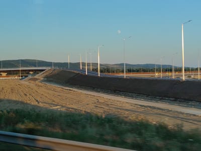 The image captures a view of an under-construction highway and bridge, with the moon visible in the clear, pale blue sky. The landscape is characterized by rolling hills covered in vegetation and trees in the background. The foreground shows a dirt road with tire tracks, suggesting recent activity, and a blurred green area at the very bottom, possibly indicating foliage seen from a moving vehicle. A metal guardrail is also visible at the bottom of the frame. Numerous streetlights line the highway, some of which are illuminated, casting a warm glow. The overall scene appears to be a semi-rural or developing area. The specific location is identified as Nicolae Bălcescu, Romania.