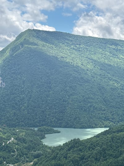 The image shows a panoramic view of a mountainous landscape in the city of Gagulechi, Georgia. The foreground features a lush, densely forested mountain slope descending towards a tranquil, light-colored body of water, likely a lake or reservoir. The water's surface reflects the sky and is bordered by the verdant trees. A winding road is visible on the left side of the frame, carving its way through the wooded terrain. In the distance, a large, imposing mountain covered in dense green forest dominates the background. The summit of this mountain is adorned with what appear to be three tall, slender structures, possibly monuments or communication towers. The sky above is a mix of blue with scattered white clouds.