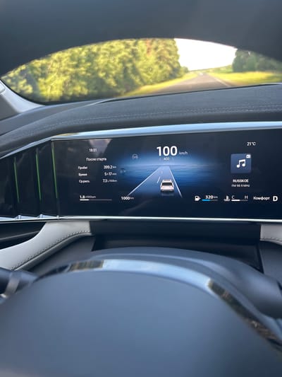 The image shows the interior of a car, looking out through the windshield and at the dashboard display. The dashboard displays various information including speed, time, mileage, fuel range, and radio station. The car is on a road with trees lining both sides, suggesting a rural or suburban environment. The time displayed on the dashboard is 18:51, and the outside temperature is 21°C. The speed is 100 km/h, and the ACC (Adaptive Cruise Control) is active. The radio is tuned to "RUSSKOE" at FM 98.9 MHz. The fuel range is 320 km. The car's interior appears modern, with a large digital display. The image does not contain any information that would allow for the determination of its location in the city of Тарноўскі сельскі Савет, Belarus.
