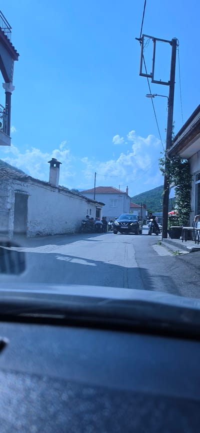 The image depicts a street scene in the city of Melivoia, Greece, captured from the interior of a vehicle, with the dashboard visible in the foreground. The sky is clear and blue with scattered white cumulus clouds.
On the left side of the street, an old, whitewashed building with a prominent chimney and a dark metal door is visible. Several motorcycles are parked against this building, and a group of approximately four individuals are gathered nearby, appearing to socialize. Further down the street, another light-colored building with visible windows stands.
On the right side of the street, a wooden utility pole equipped with electrical wires, a street light, and a distinctive rectangular wooden frame structure is present. Adjacent to the pole is a building adorned with climbing greenery, featuring outdoor seating with chairs and a partially visible red awning, suggesting a commercial establishment. A man is seated on a motorcycle on the right side of the road, facing towards the camera.
In the center of the asphalt road, a dark-colored Nissan Qashqai SUV with the license plate "ETE 0293" is approaching the viewer. Shadows are cast onto the road surface in the immediate foreground. In the background, distant hills or mountains are visible under the clear sky.