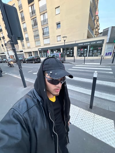 The image displays an urban street scene in Bois-Colombes, France, captured from a low perspective. In the immediate foreground, positioned to the lower right, is a male individual with light skin and facial stubble. He is attired in a black baseball cap featuring metallic studs and rings, dark wraparound sunglasses, a black hooded jacket with the hood raised, a black t-shirt, and an open black leather-like jacket over the hoody. A yellow garment collar is visible beneath the black t-shirt. The individual is looking slightly downwards and to the left. He stands on a section of sidewalk with tactile paving, distinguished by raised white circular patterns.
Adjacent to the individual on the left is a dark grey pole, likely a street sign support, with a circular sign affixed. The street itself contains a pedestrian crossing marked with white zebra stripes. On the left side of the street, several cars are parked, and a scooter or motorcycle is in motion, moving away from the camera. A black bollard with a white top delineates the pedestrian crossing on the right side of the street, near a modern black street lamp.
The background features a multi-story, light-colored building with multiple windows and balconies, suggesting a residential or mixed-use structure. The ground floor accommodates a commercial establishment identified as a real estate agency by the prominent sign "VENDRE ACHETER LOUER GÉRER" and "lamy" displayed across its windows, which exhibit various property listings. The sky above is clear and light blue, indicating daylight.